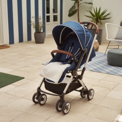 Modern blue stroller with a leather-accented handle and cozy seat, accessorized with a soft white blanket, set on a sunny patio.