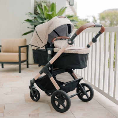 Elegant beige bassinet-style stroller with a textured canopy and leather-accented handle, perfect for comfort and style on a breezy veranda.