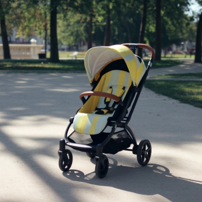 A stylish yellow and white striped baby stroller with a sun canopy, positioned on a sunlit park pathway with lush green trees and a water fountain in the background.