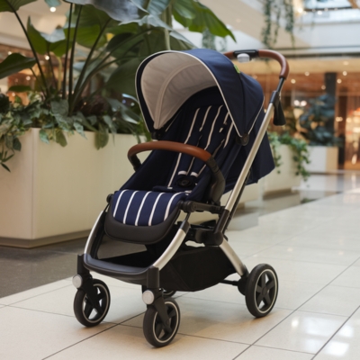 Navy blue and white striped baby stroller with a sturdy black frame and brown handlebar, parked in a modern shopping mall near lush greenery, designed for different weight limits and age stages.