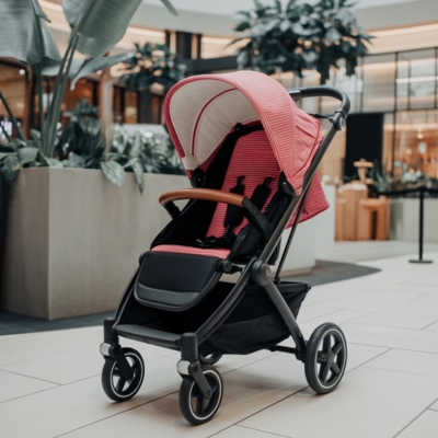 Red and black baby stroller with a striped canopy and brown handlebar, parked in a shopping mall near lush indoor plants, offering a stylish and space-efficient design for small homes.